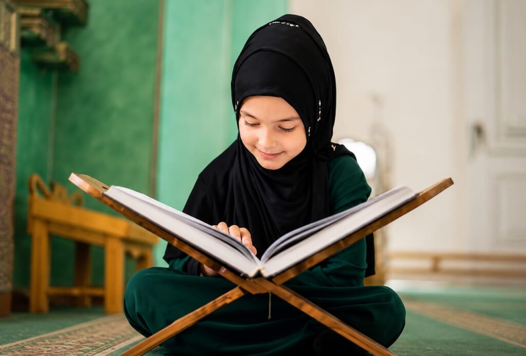 A young Muslim girl wearing a black hijab sitting cross-legged and smiling while reading the Holy Quran on a wooden stand.